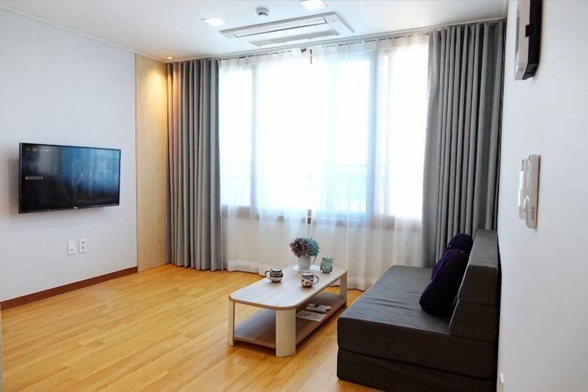 Una sala de estar con televisor de pantalla plana, un sofá, una mesa de centro y una ventana con cortinas.