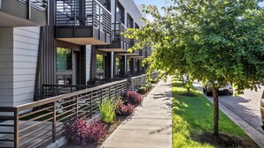 Un edificio moderno de varios pisos con balcones, una acera y un árbol.
