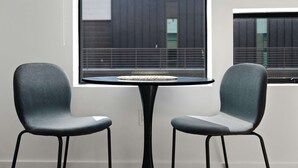 Una habitación con dos sillas y una mesita, una ventana que da a un edificio, y una pared blanca.