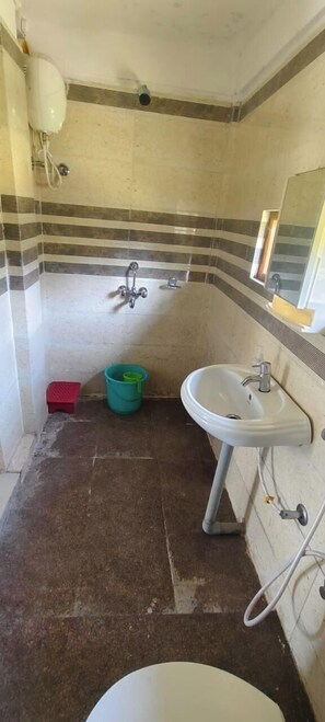 A small bathroom with a white sink, a mirror, and a shower. There is a red stool and a green bucket on the floor. - Kinglayang Homestay (Kalimpong)