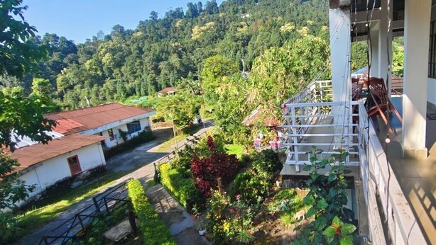 A balcony with a view of a lush green hillside, a white building with a red door, and a blue bucket on the ground. - Kinglayang Homestay (Kalimpong)