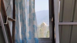A small, rustic room with a wooden floor, a window with a blue and white curtain, and a view of the outdoors. - Kinglayang Homestay (Kalimpong)