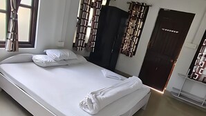 A neatly made bed with white linens and pillows, a dark-colored wardrobe, and a door with a patterned curtain. - Kinglayang Homestay (Kalimpong)