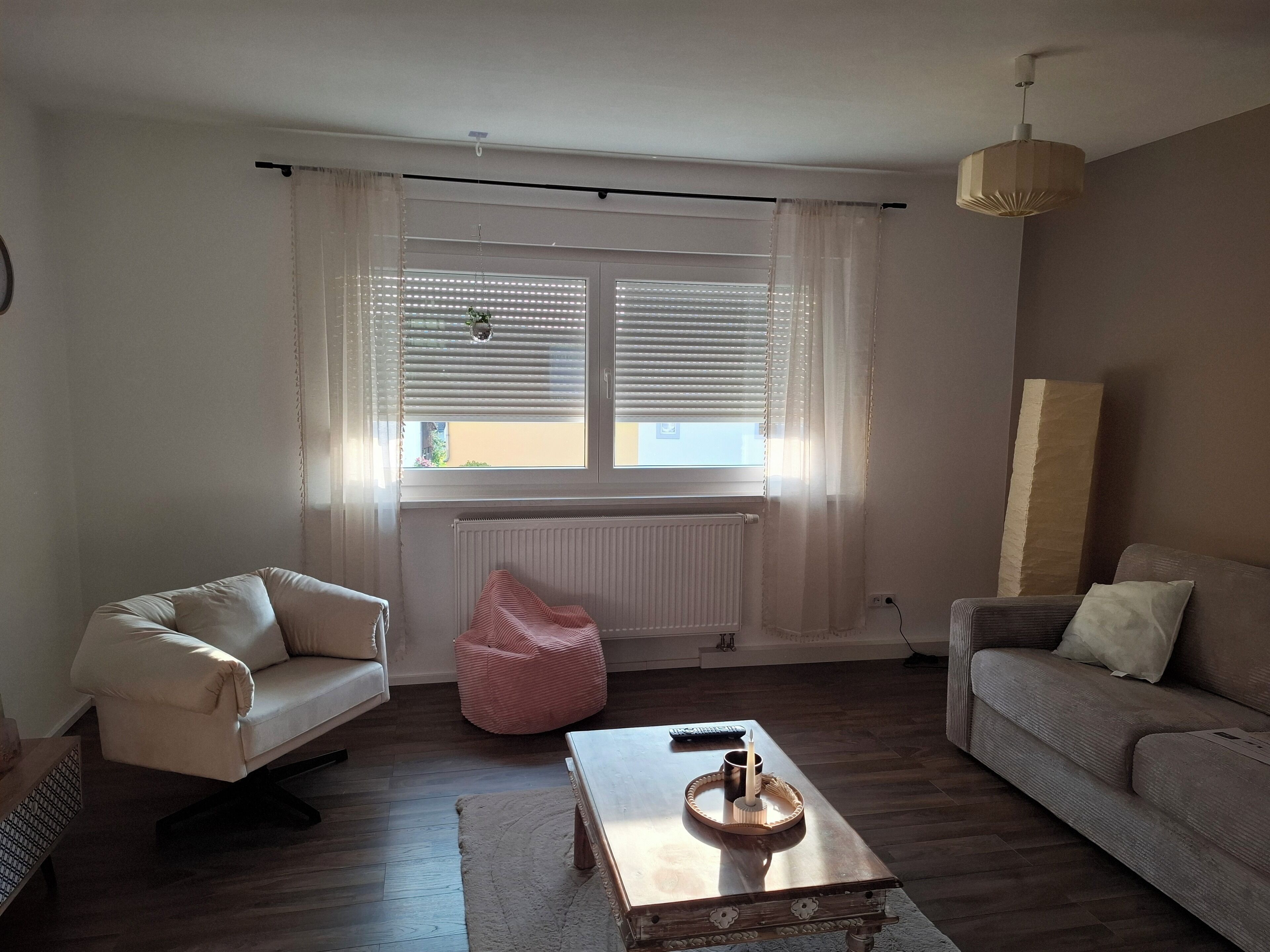Een woonkamer met een bank, een stoel, een bijzettafeltje met een kaars en dienblad, een radiator, en een raam met zonwering.
