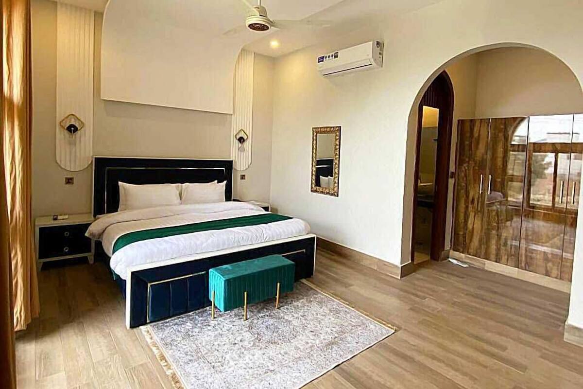 A bedroom with a bed, bedside tables, a ceiling fan, an air conditioning unit, a mirror, and a view of the outside through an arched doorway.