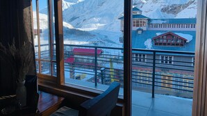 A room with a large window overlooking a snowy mountain landscape and a small building with a blue roof. - The Bulevar-Whispering Willow (Lahul)
