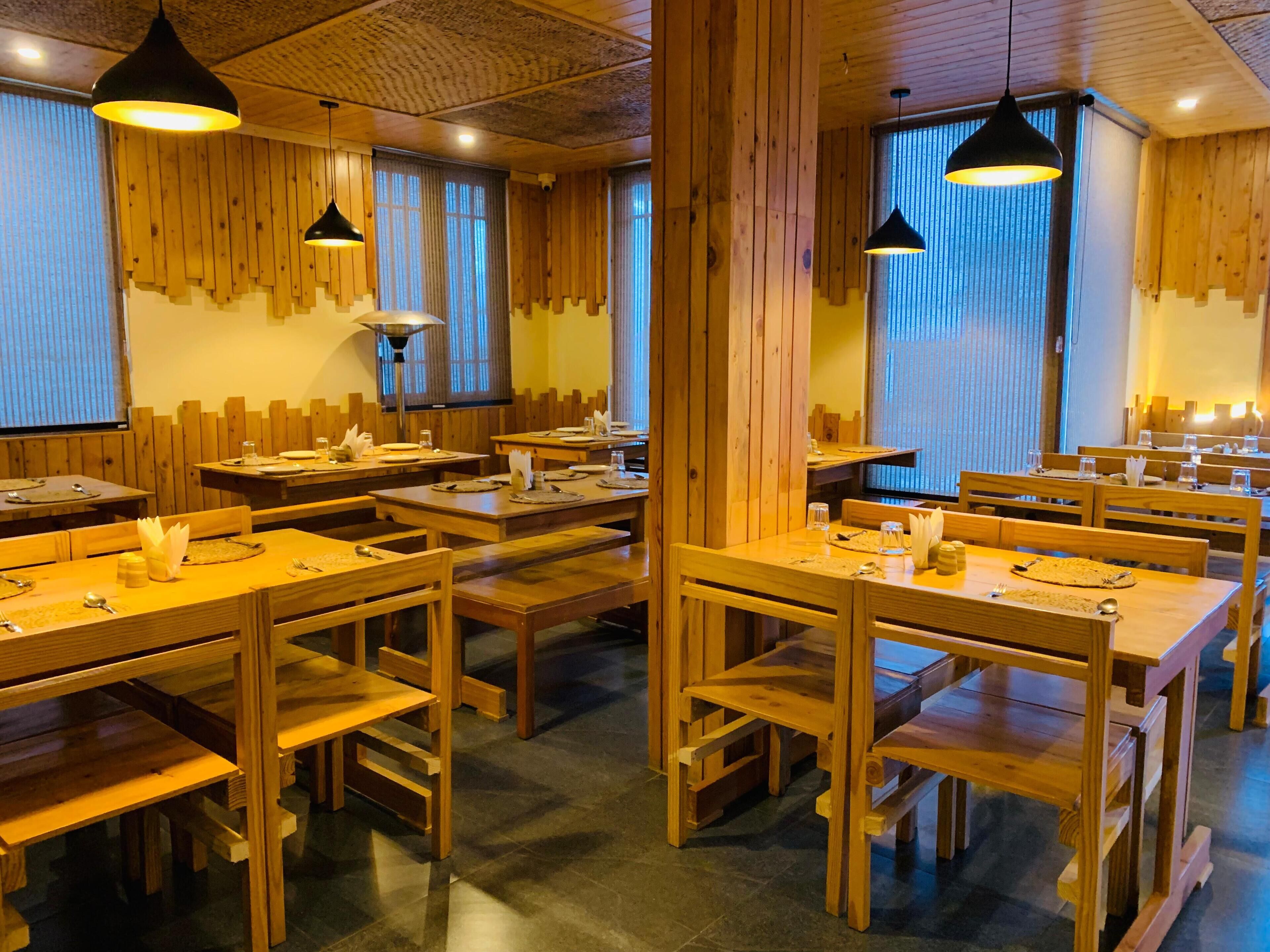 Un restaurant avec des tables et des bancs en bois, des luminaires suspendus et de grandes fenêtres ornées de rideaux.