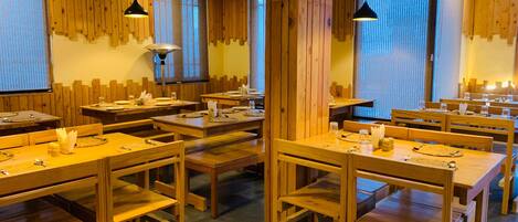 Un restaurant avec des tables et des bancs en bois, des luminaires suspendus et de grandes fenêtres ornées de rideaux.