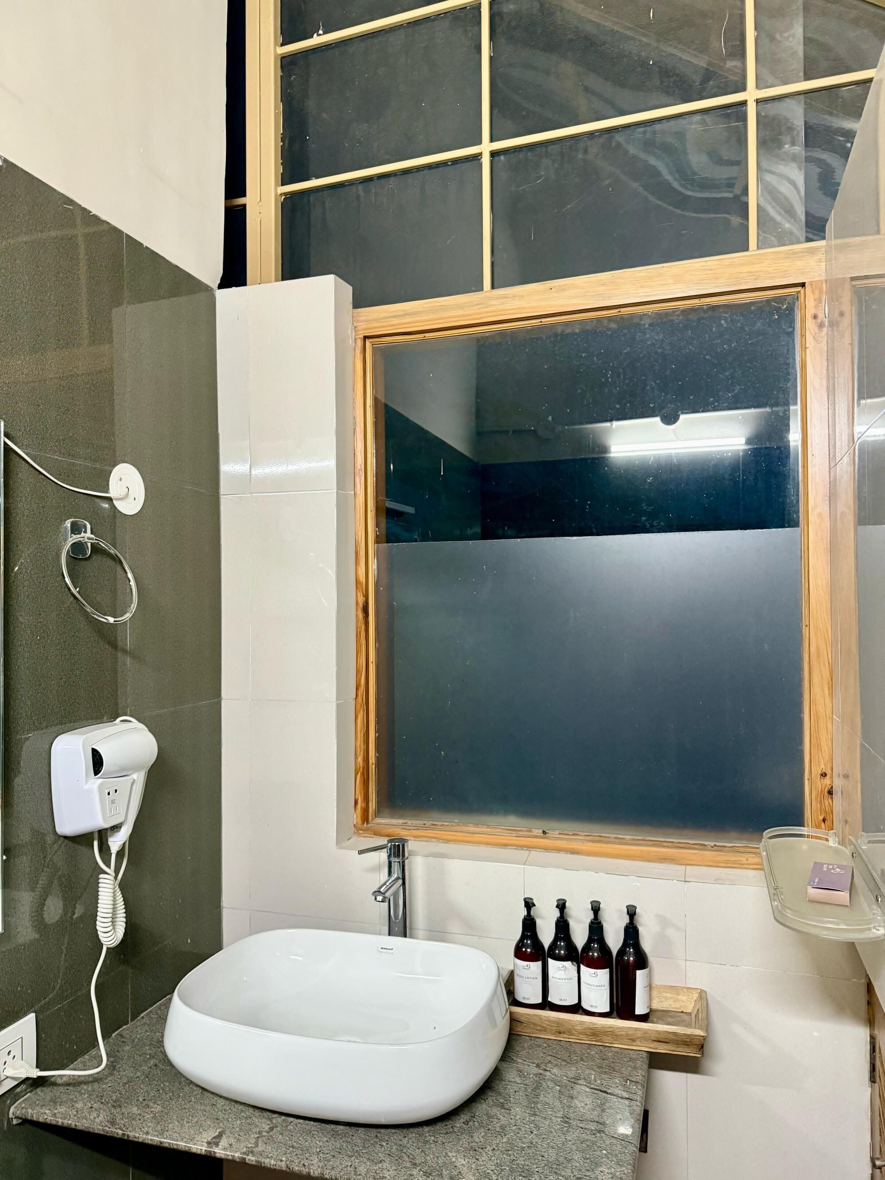 Un baño con lavamanos blanco, secador de pelo y ventana con marco de madera.