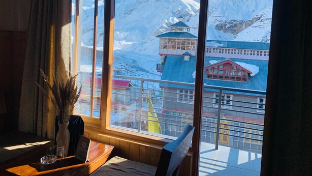 A room with a large window offering a view of snow-covered mountains and a building with a blue roof. - The Bulevar-Whispering Willow (Lahul)
