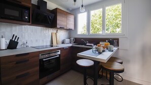 A modern kitchen with wooden cabinets, a microwave, a stove, and a sink. There are stools at the island and a window with a view of greenery. - Le Mathurin - Wonderful 4 rooms. (Saint-Brieuc)