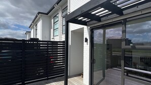 Ein modernes Haus mit einer Holzdielen-Terrasse, einem schwarzen Zaun und einer Glastür, die zu einem Außenbereich führt.