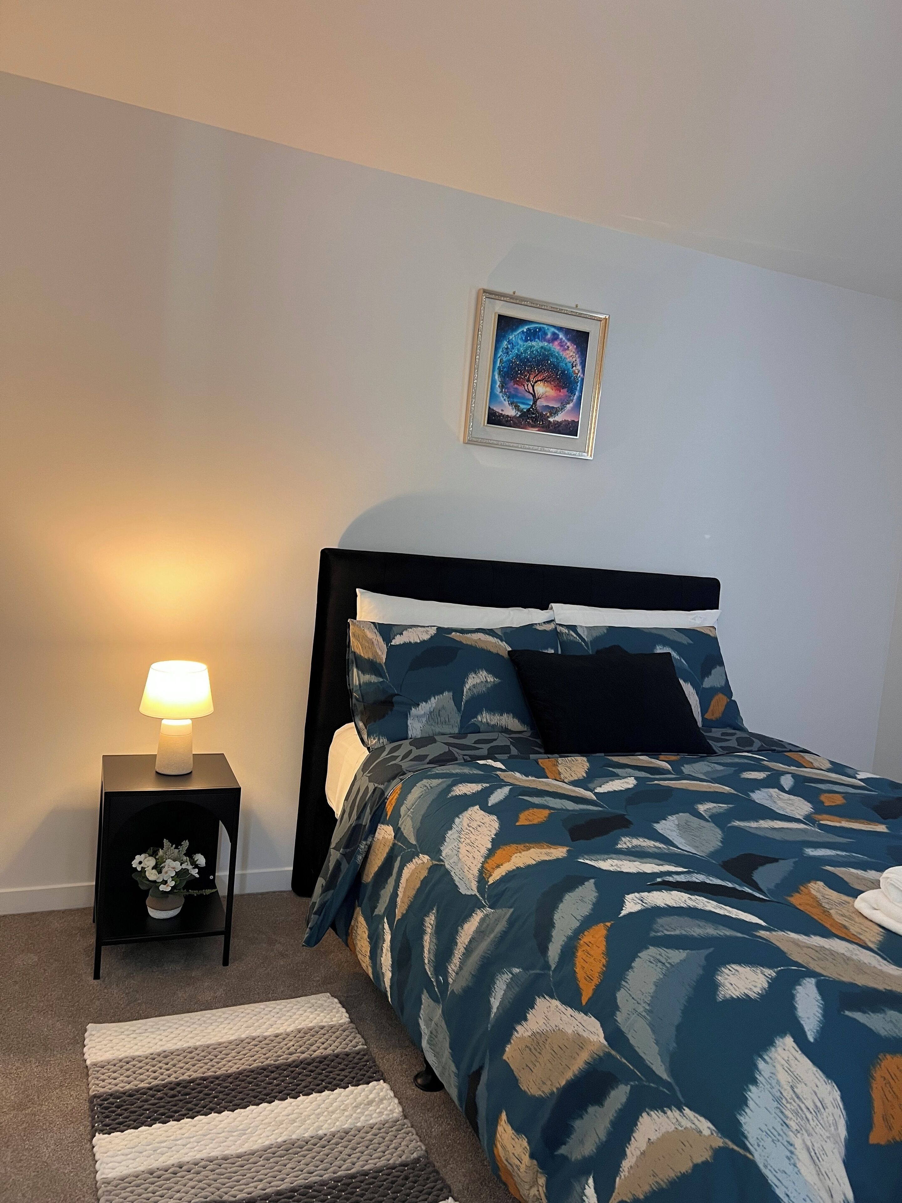 Un dormitorio con cama, mesita de noche con lámpara, un cuadro en la pared y una alfombra.
