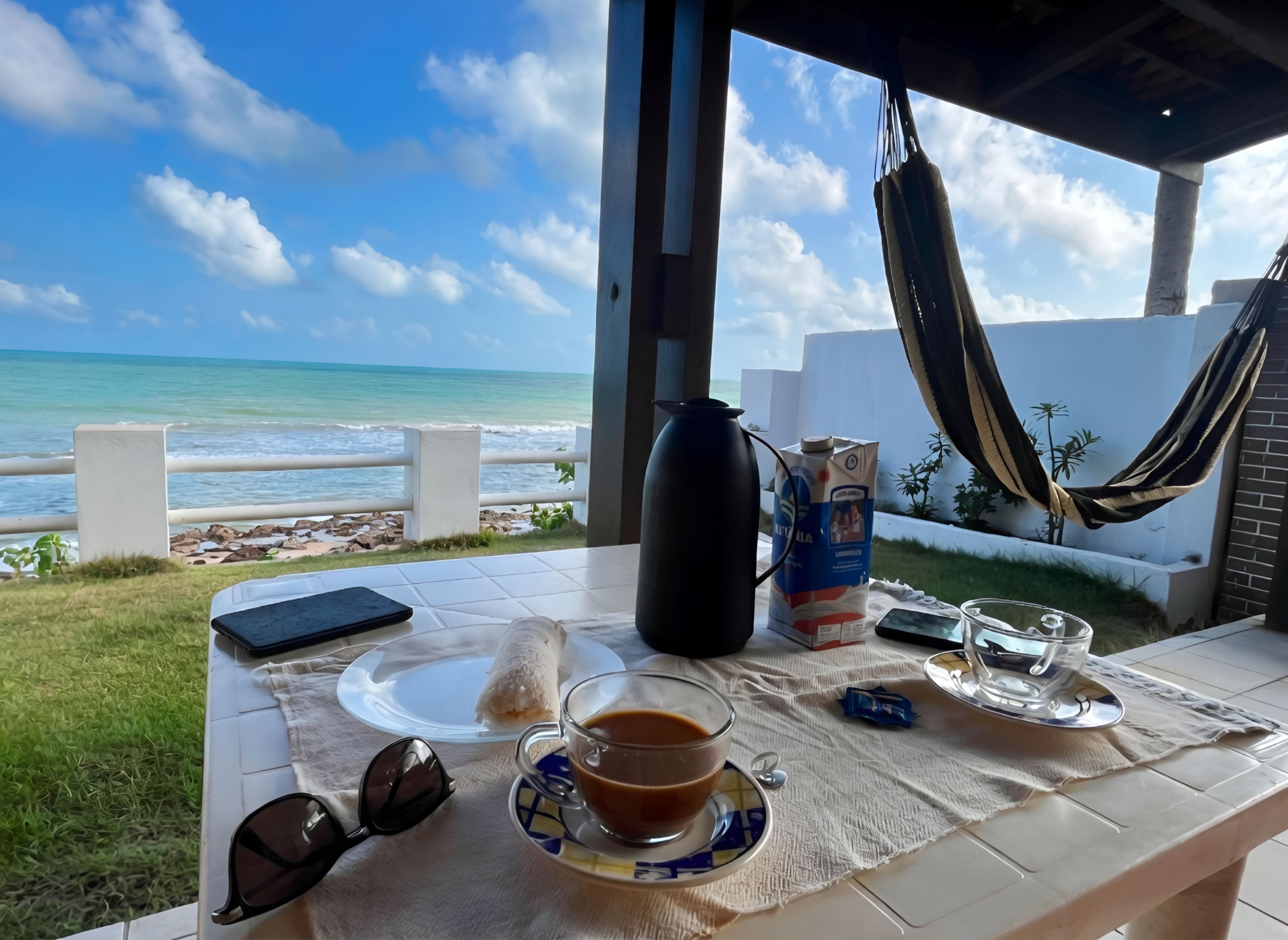 Una mesa con una taza de café, un plato de comida, gafas de sol, un teléfono móvil y una hamaca con vista a una playa.