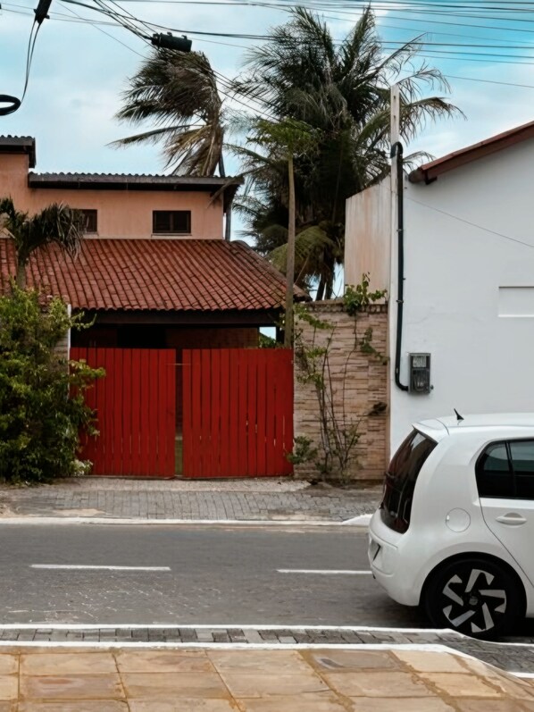 A street view with a red gate, a white car parked on the side, and a building with a tiled roof.