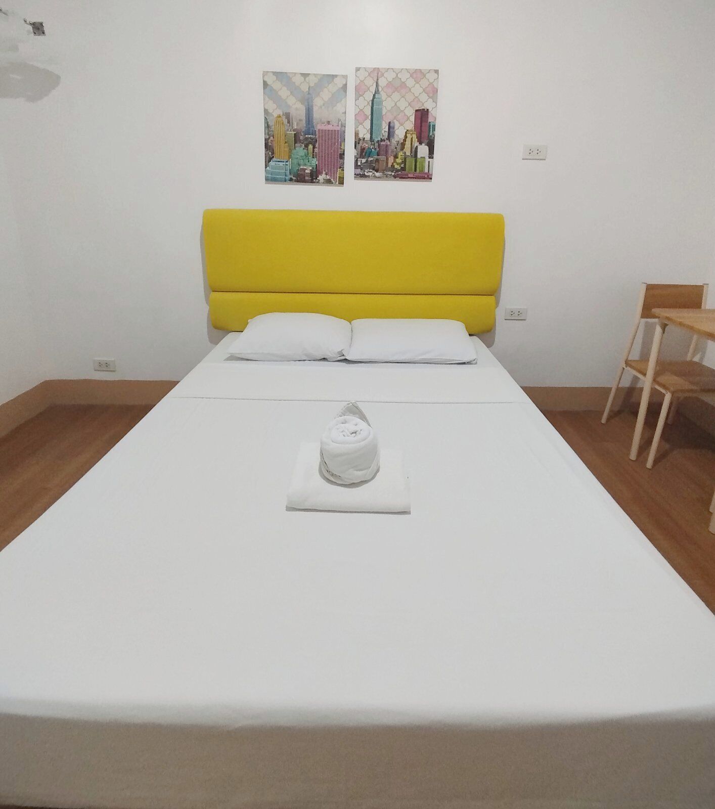 Un lit bien fait, avec une tête de lit jaune, du linge de lit blanc et une serviette enroulée posée sur un oreiller blanc. Deux œuvres d’art sont accrochées au mur et représentent des paysages urbains.
