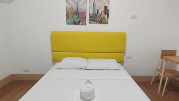Un lit bien fait, avec une tête de lit jaune, du linge de lit blanc et une serviette enroulée posée sur un oreiller blanc. Deux œuvres d’art sont accrochées au mur et représentent des paysages urbains.