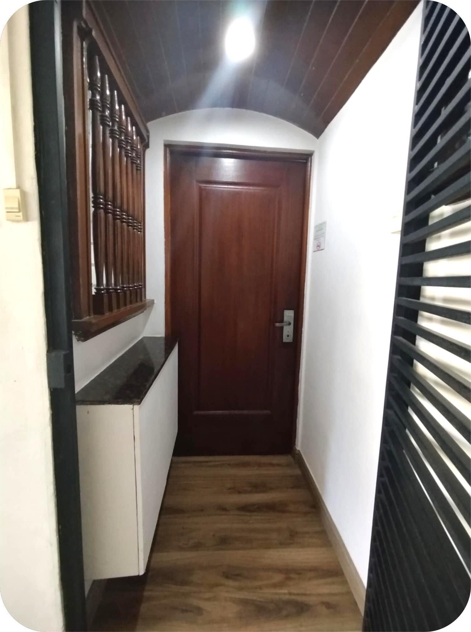 A narrow hallway with a wooden door at the end, a window with blinds on the right, and a light fixture on the ceiling.