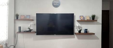 A living room with a flat-screen TV mounted on the wall, a ceiling fan, and wooden shelves with decorative items.