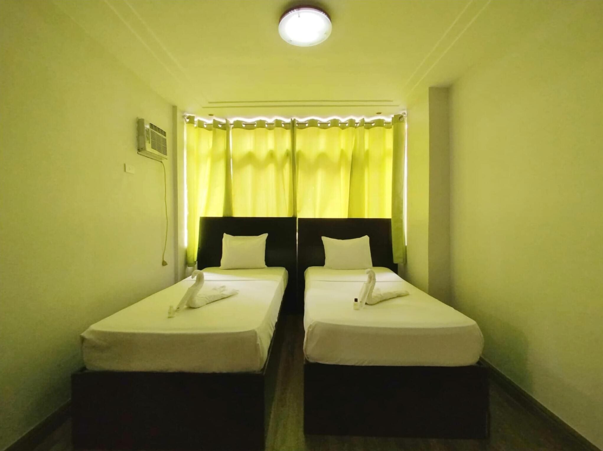 A hotel room with two single beds, a headboard, a window with yellow curtains, and a ceiling light.
