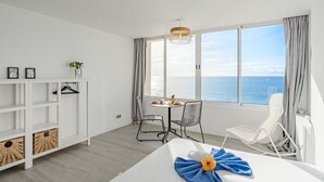 A modern hotel room with a large window offering a sea view, a dining area with a table and chairs, and a white bed with a blue towel and flower. - Ocean Loka by Interhome (Callao Salvaje)