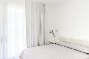 A minimalist bedroom with a large bed, a nightstand, a vase with a plant, and sheer curtains.