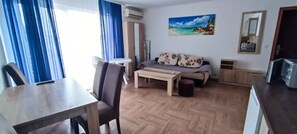 Een woonkamer met een bank, een salontafel, een eettafel met stoelen, een televisie en een schilderij aan de muur.