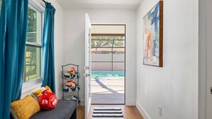 Um corredor com vista para uma piscina, um banco com almofadas, uma estante de metal com roupas dobradas e um quadro na parede.