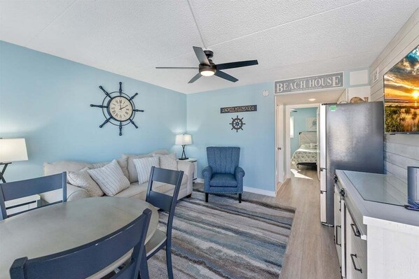 A room with a dining table, chairs, a blue armchair, a ceiling fan, a clock, and a kitchen area with a refrigerator.