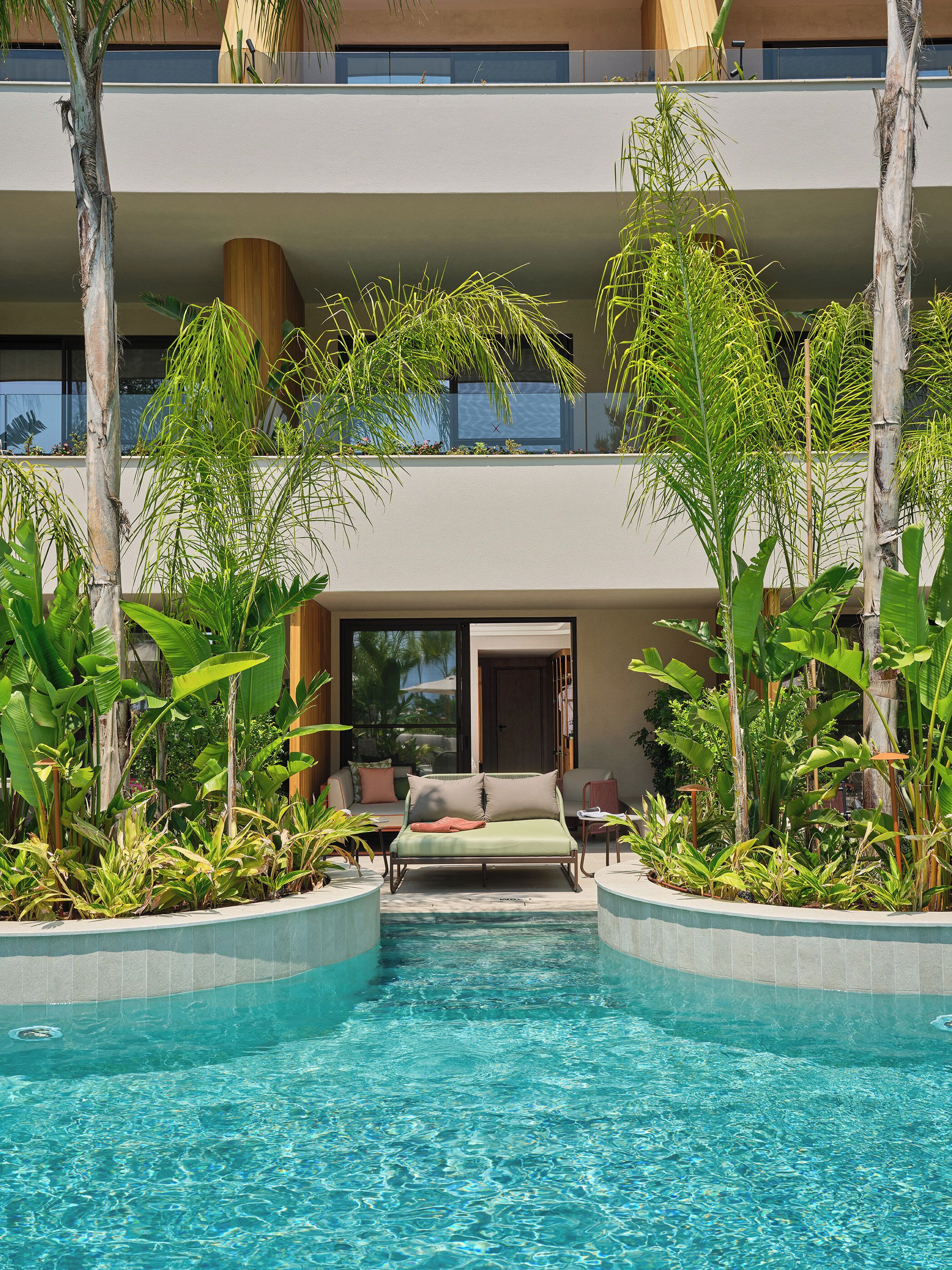 Un área de piscina con una cama y sillas de descanso, rodeada de plantas tropicales.