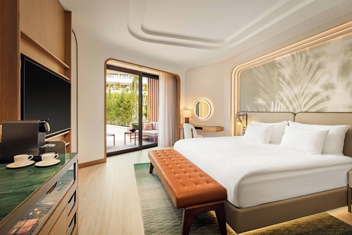 A modern hotel room with a large bed, a desk with a laptop, a coffee station, and a view of the outdoors.