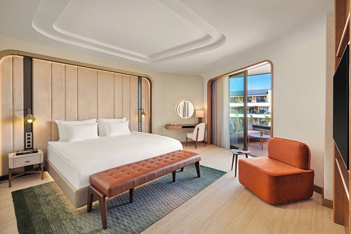 A modern hotel room with a large bed, a seating area with a bench and armchair, a desk with a chair, and a large window offering a view of the city.