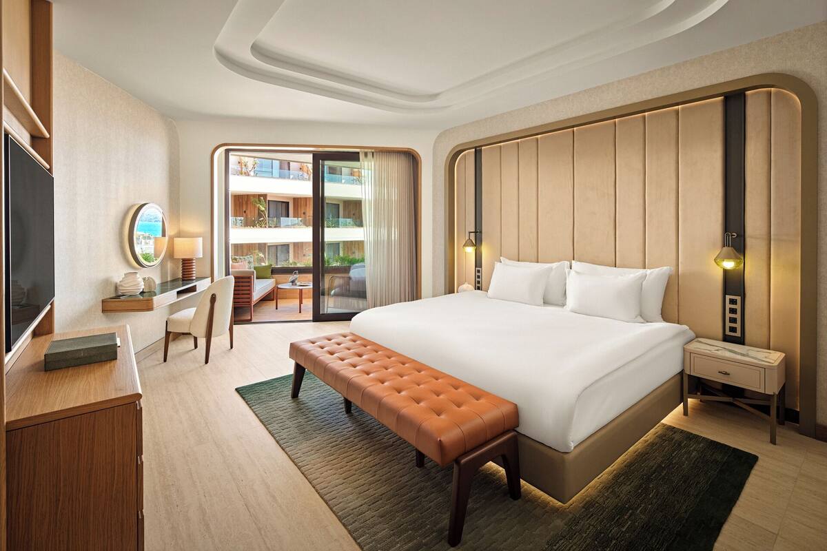 A hotel room with a large bed, a desk with a chair, a bench, a TV, and a view of the outside balcony.
