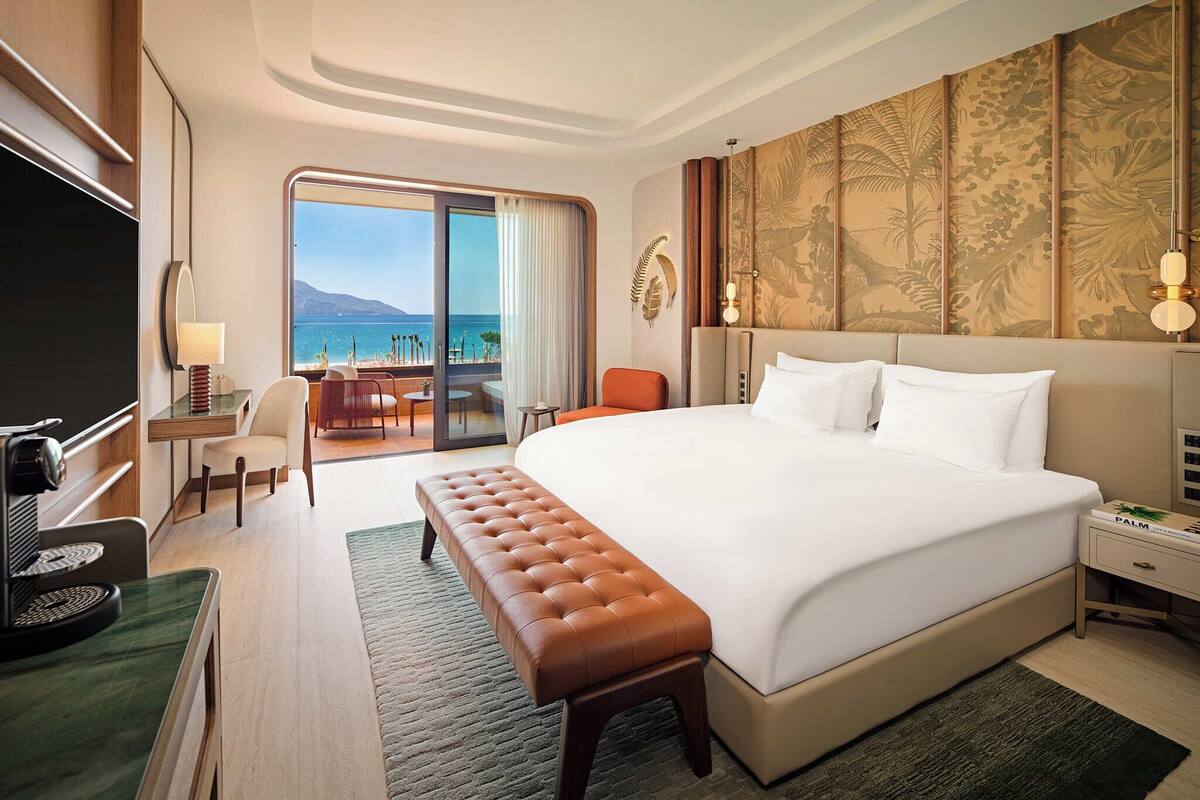 A hotel room with a large bed, a desk, a chair, a TV, and a balcony with a view of the sea.