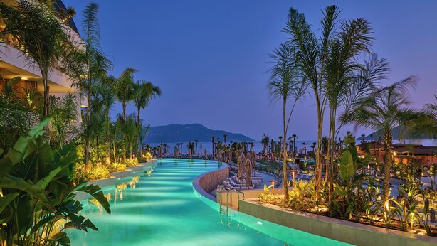 A resort pool area with a curved design, surrounded by palm trees and lounge chairs, with a view of the ocean and mountains in the background. - XO Cape Arnna Fethiye (Fethiye)