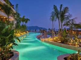 Zona de piscina en un resort con diseño curvo, rodeada de palmeras y sillas de descanso, con vistas al océano y montañas al fondo.