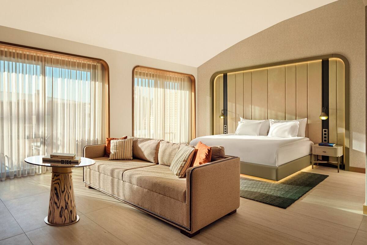 A modern hotel room with a large bed, a sofa, a small table, and large windows with curtains.