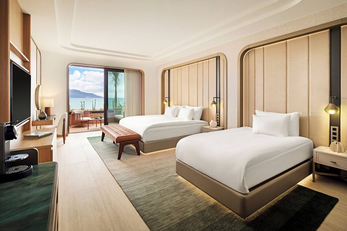 A hotel room with two beds, a desk, a chair, a TV, and a view of the sea.