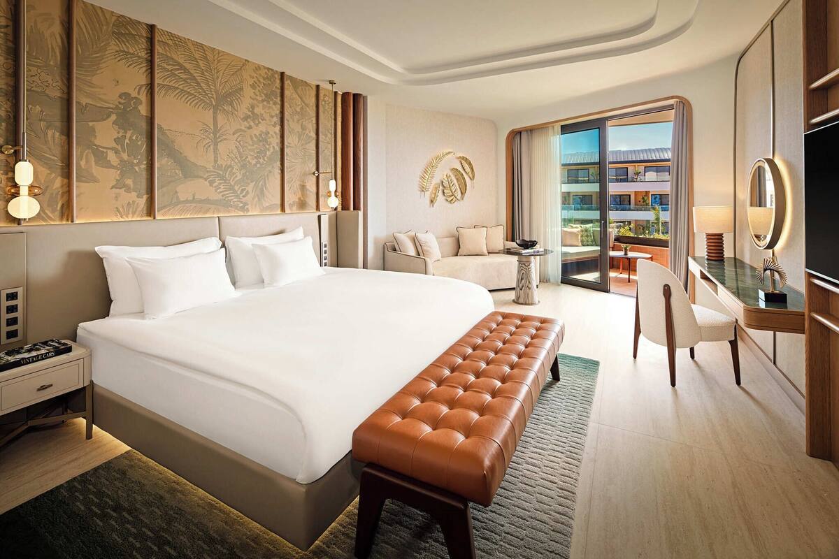 A hotel room with a large bed, a seating area with a sofa and armchairs, a desk with a mirror, and a view of the outdoor balcony.