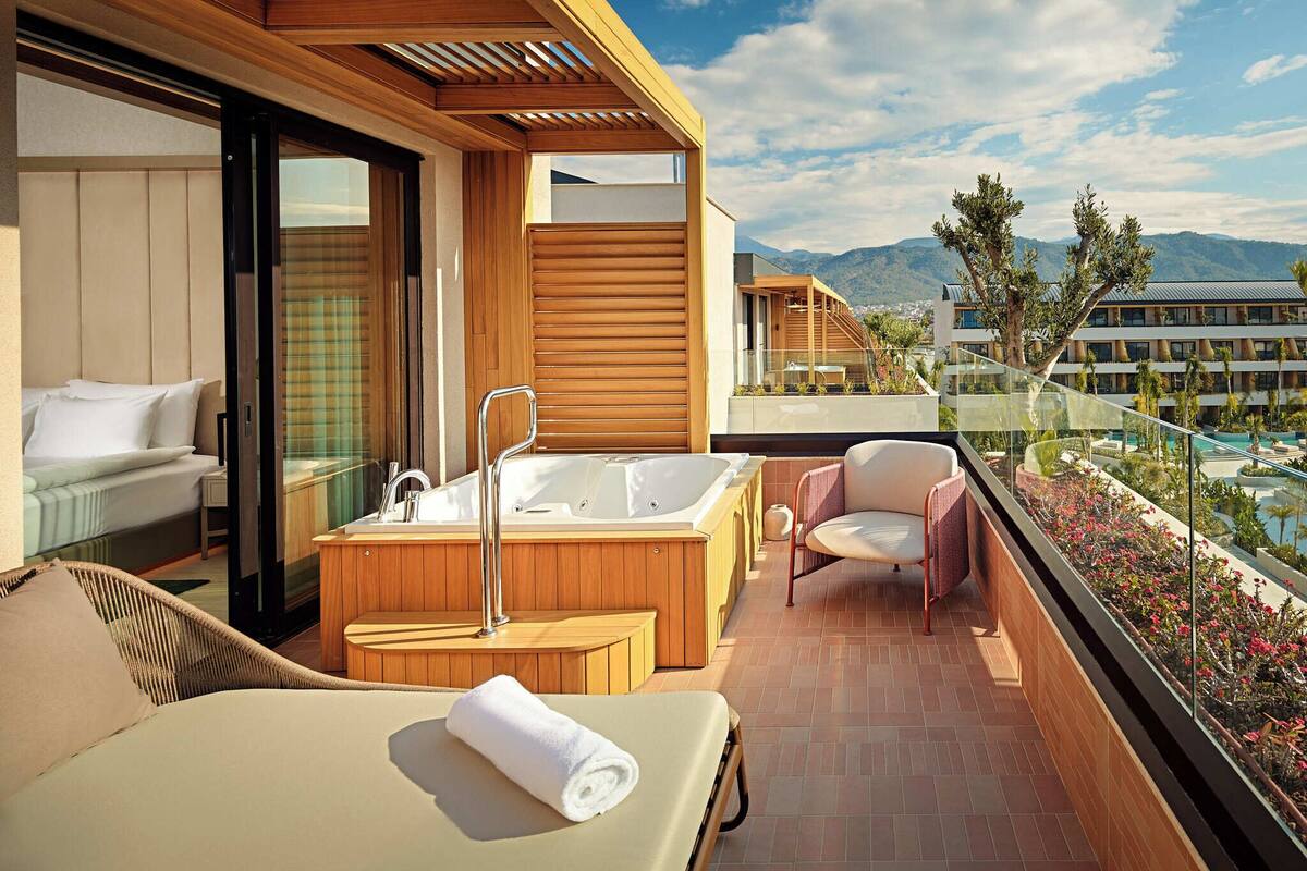 A balcony with a bed, a bathtub, and a chair, overlooking a pool and mountains.