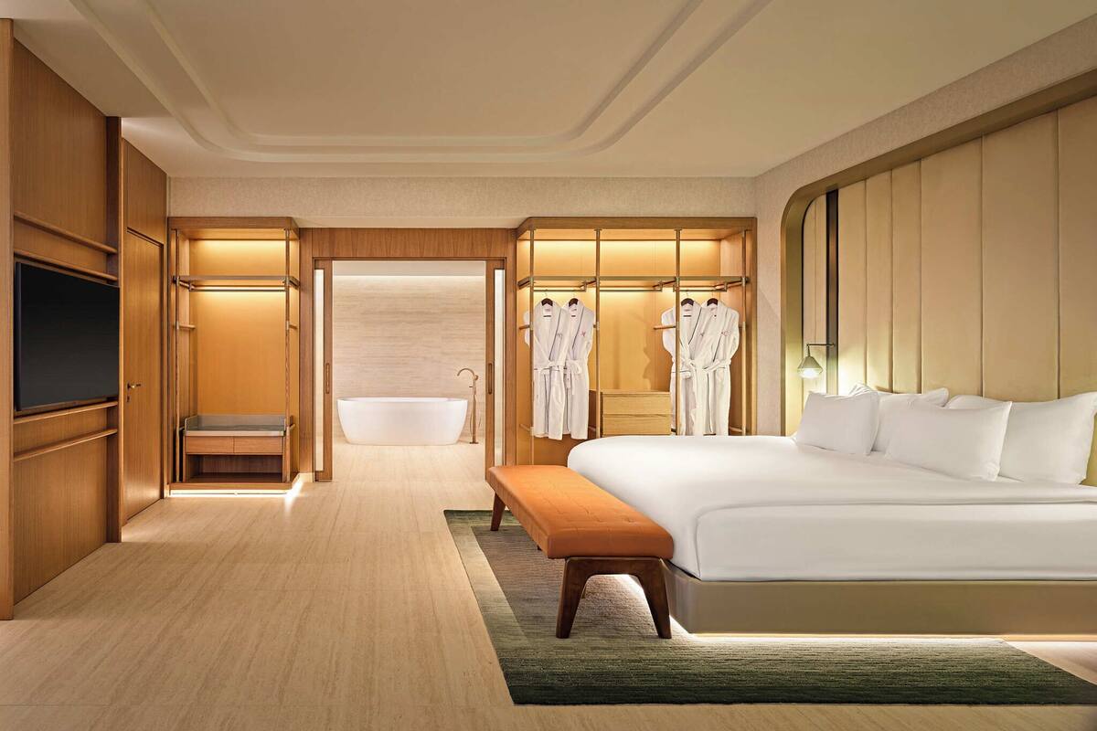 A modern hotel room with a large bed, a bathtub, a vanity with a mirror, and a walk-in closet with robes.