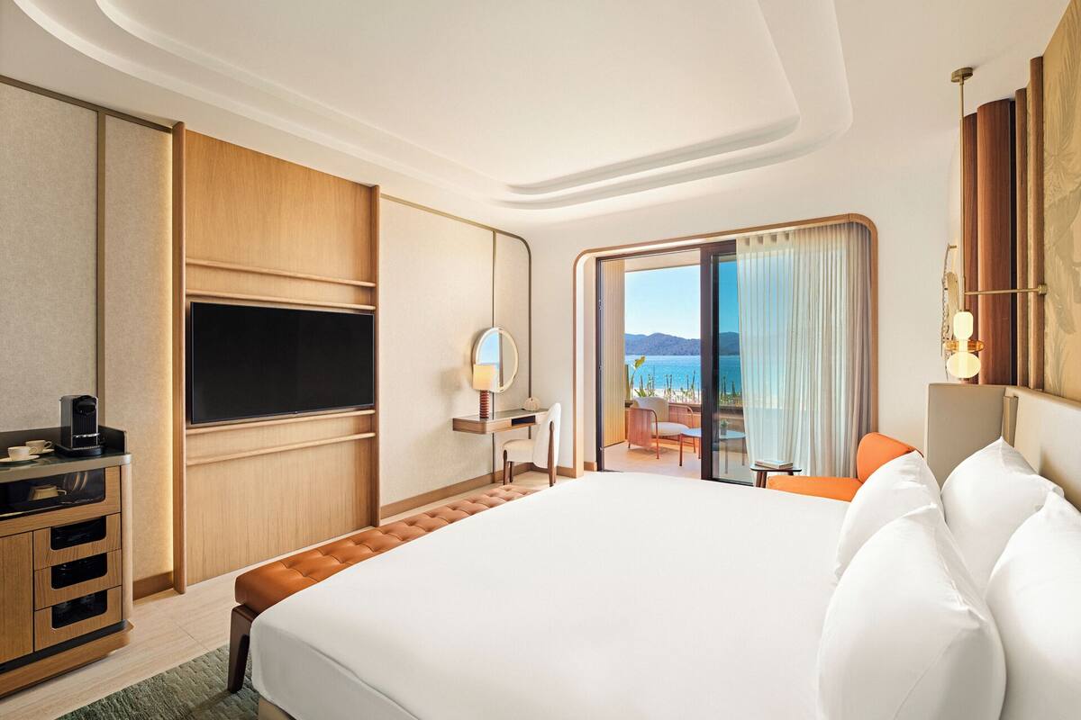 A hotel room with a large bed, a TV, a desk with a lamp, and a view of the sea.