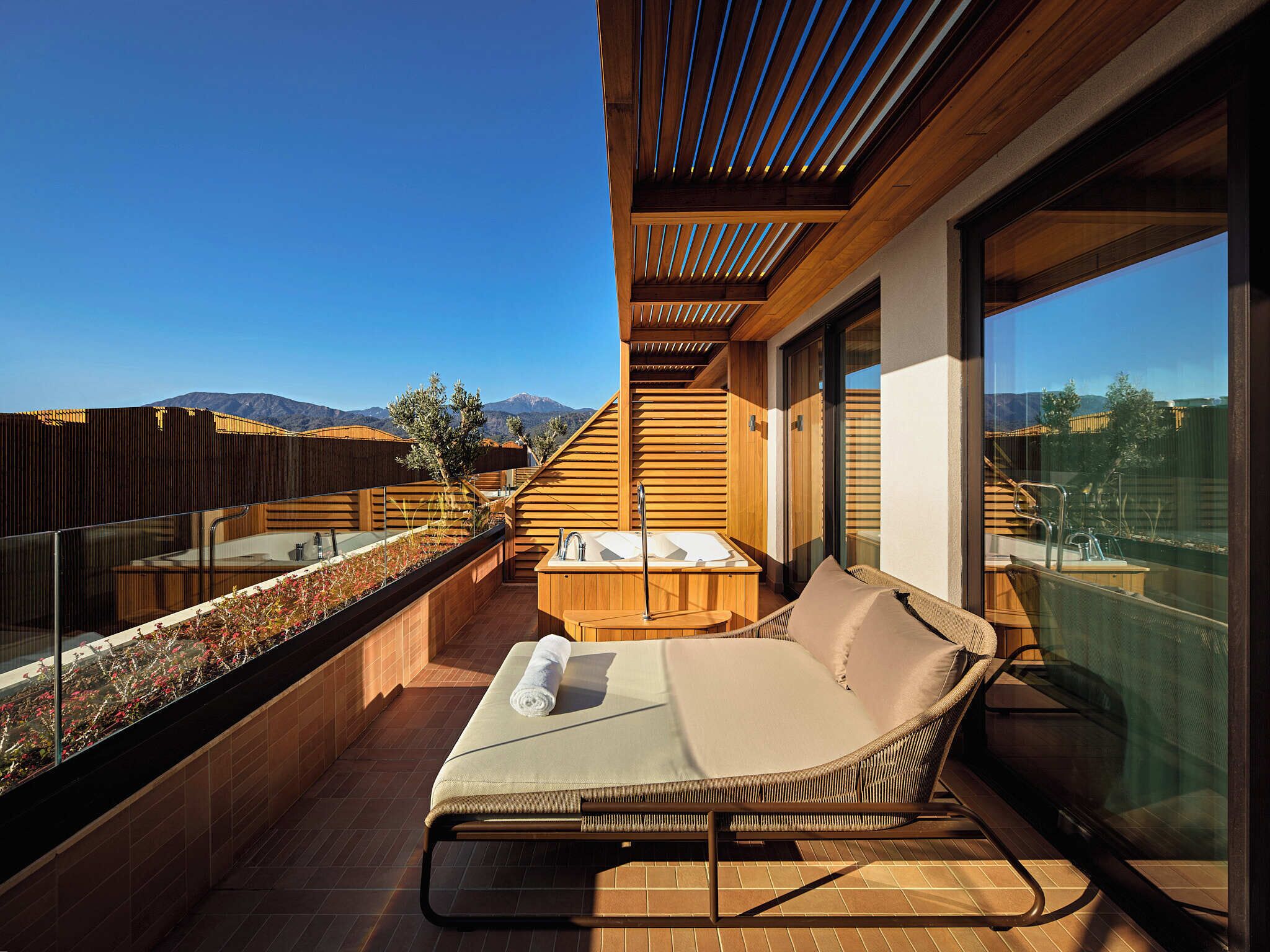 A balcony with a wooden deck, a lounge chair, a hot tub, and a glass railing overlooking a mountainous landscape.