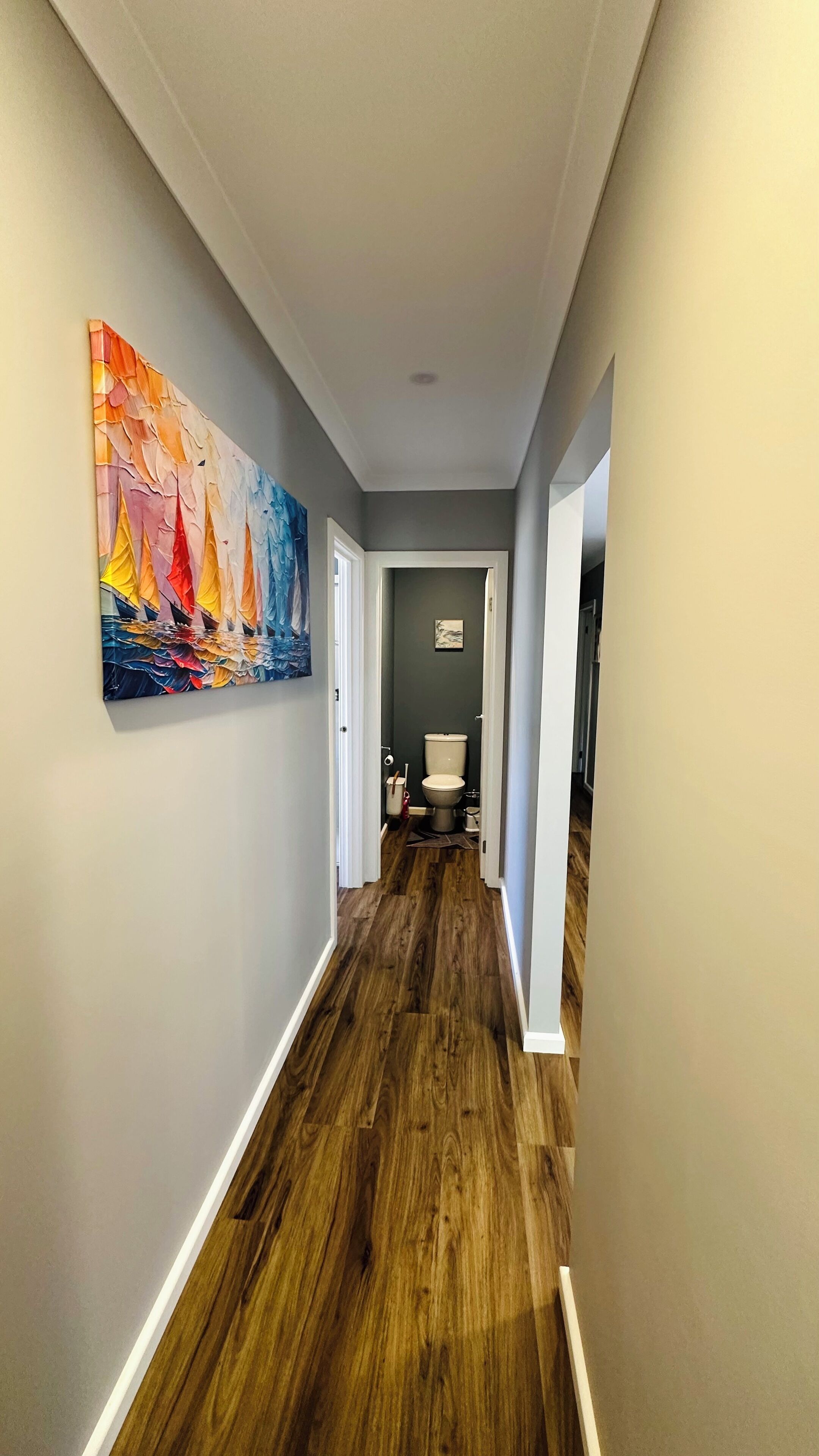 A hallway with a wooden floor, a painting on the left wall, and a bathroom visible at the end.