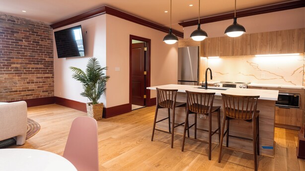A modern kitchen with a central island, wooden chairs, a flat-screen TV, and a brick wall. - Gisele Hotel (New Orleans)