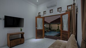 A living room with a flat-screen TV, a wooden console table, a beige sofa, and a sliding glass door leading to a balcony with a view of a building and a pool.