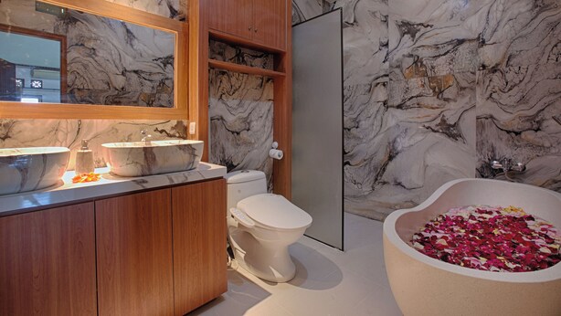 A bathroom with marble walls, a wooden vanity, a toilet, and a bathtub filled with rose petals.