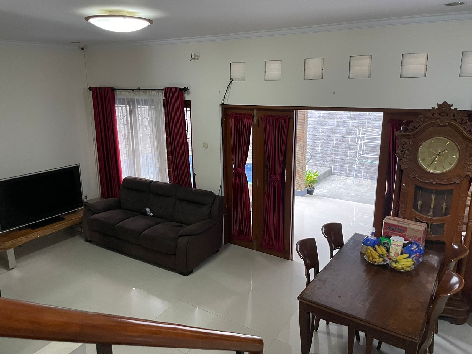 A living room with a dark sofa, a flat-screen TV, a wooden dining table with chairs, and a large wall clock.