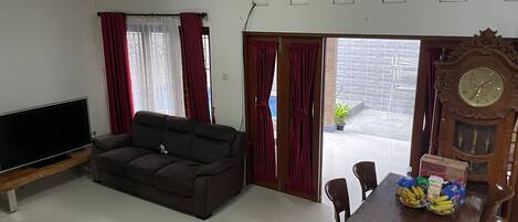 A living room with a dark sofa, a flat-screen TV, a wooden dining table with chairs, and a large wall clock.