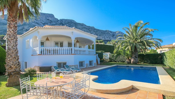 Una casa de dos pisos con piscina, mobiliario exterior y montañas al fondo.